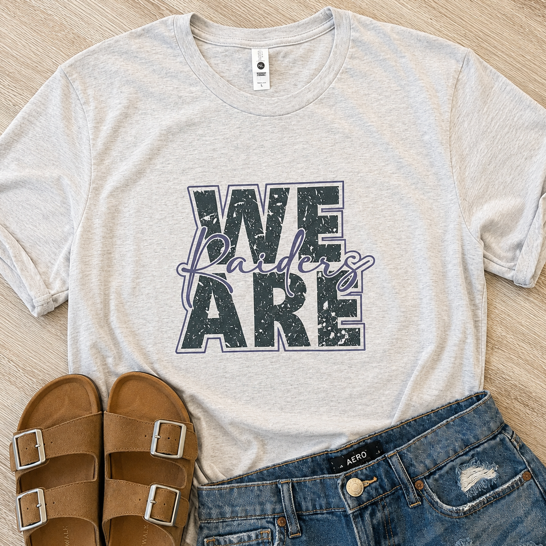 A light heather gray graphic T-shirt laid flat on a wood surface, styled with brown slide sandals and blue distressed denim shorts. The shirt has large block text reading 'WE ARE' with a purple script overlay that appears to say 'Flanders.