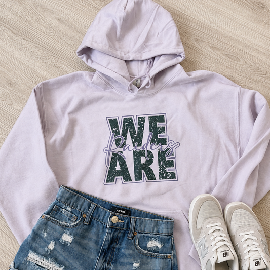 A styled flat-lay of a light lavender hoodie with a distressed green and purple 'WE ARE' graphic, paired with distressed denim shorts and white New Balance sneakers on a light wood floor.