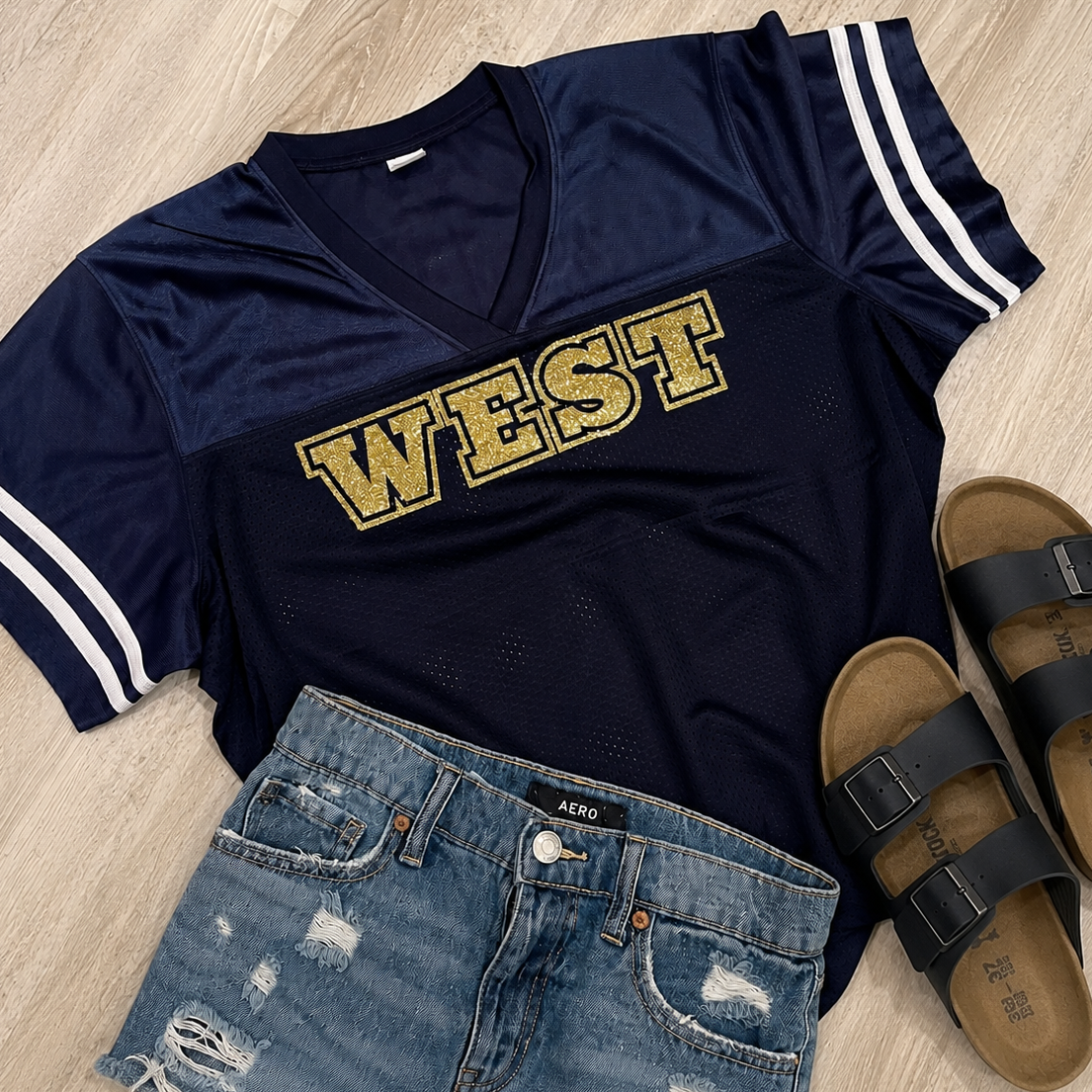 A navy blue V-neck athletic jersey top with 'WEST' in gold block letters across the chest, styled in a flat lay with distressed denim shorts, black slide sandals, and white stripe details on the sleeves, all on a light wood floor.