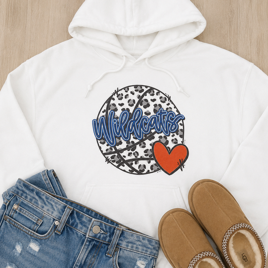 A white pullover hoodie laid flat on a light wood surface, styled with blue jeans and tan slippers. The hoodie has a front kangaroo pocket and a graphic print featuring a leopard-print basketball, blue outlined script text reading 'Wildcats,' and a red heart.