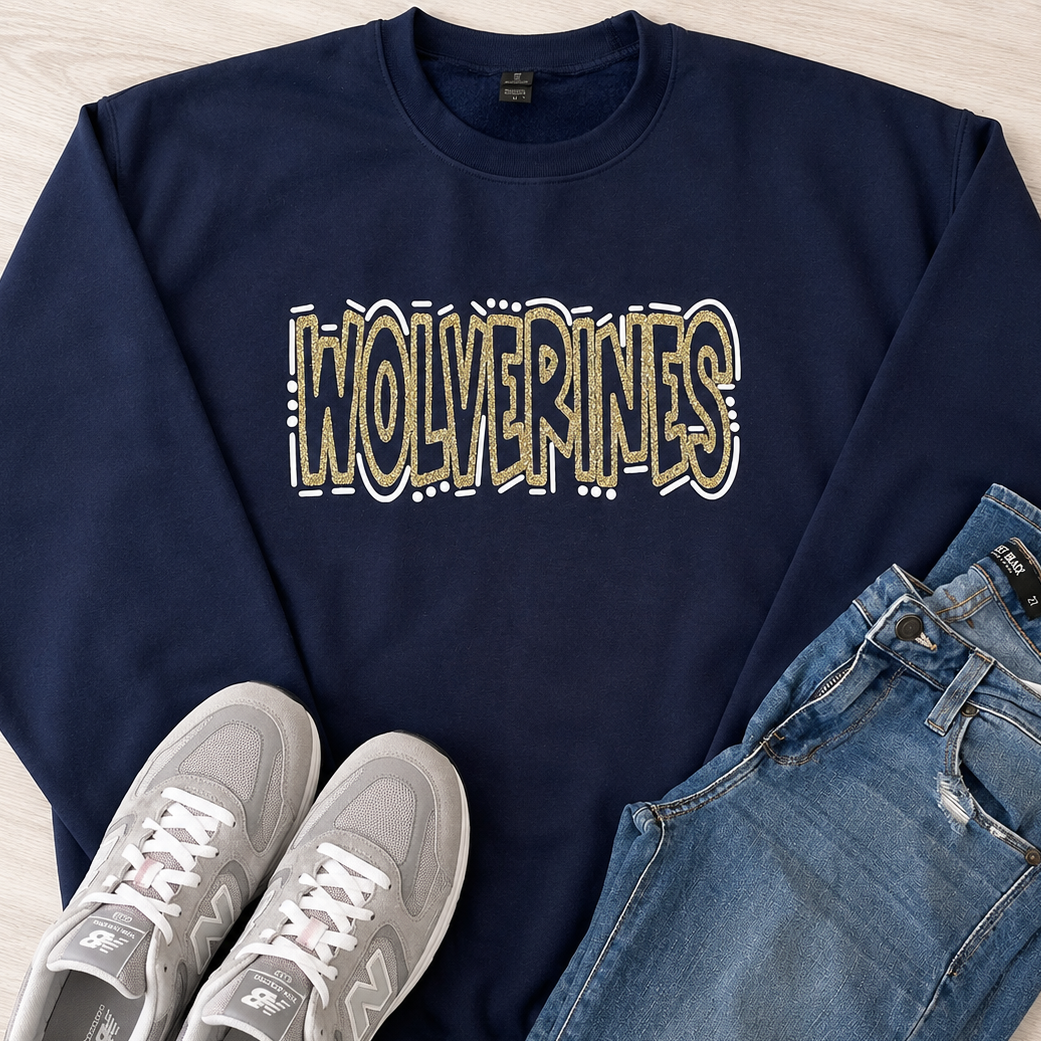 A navy blue crewneck sweatshirt with 'WOLVERINES' printed across the chest in a stylized gold and white graphic, styled in a flat lay with blue jeans and gray New Balance sneakers on a light wood floor.