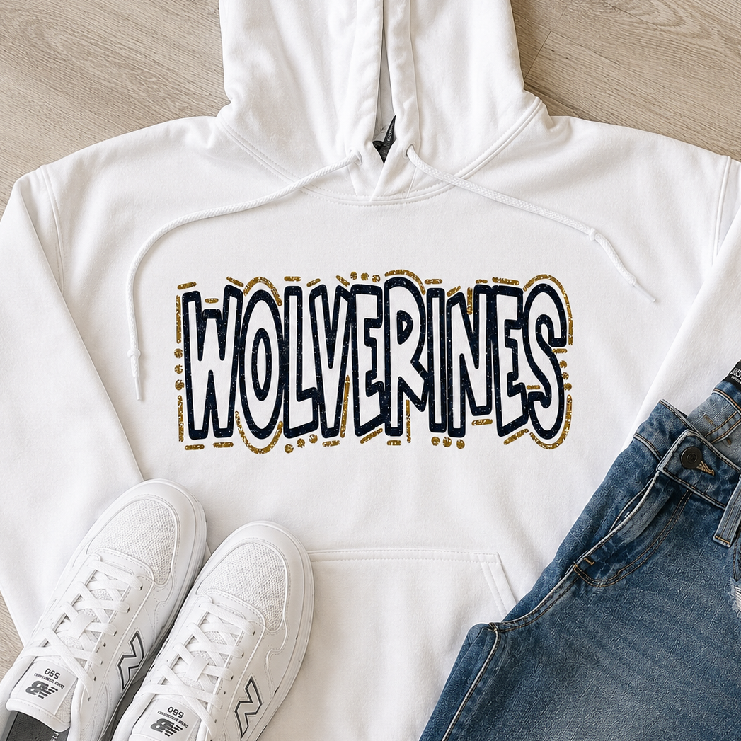 A white pullover hoodie laid flat on a light wood floor, featuring the word 'WOLVERINES' across the chest in a black, white, and gold graphic. White New Balance sneakers and blue jeans are styled around it as outfit props.