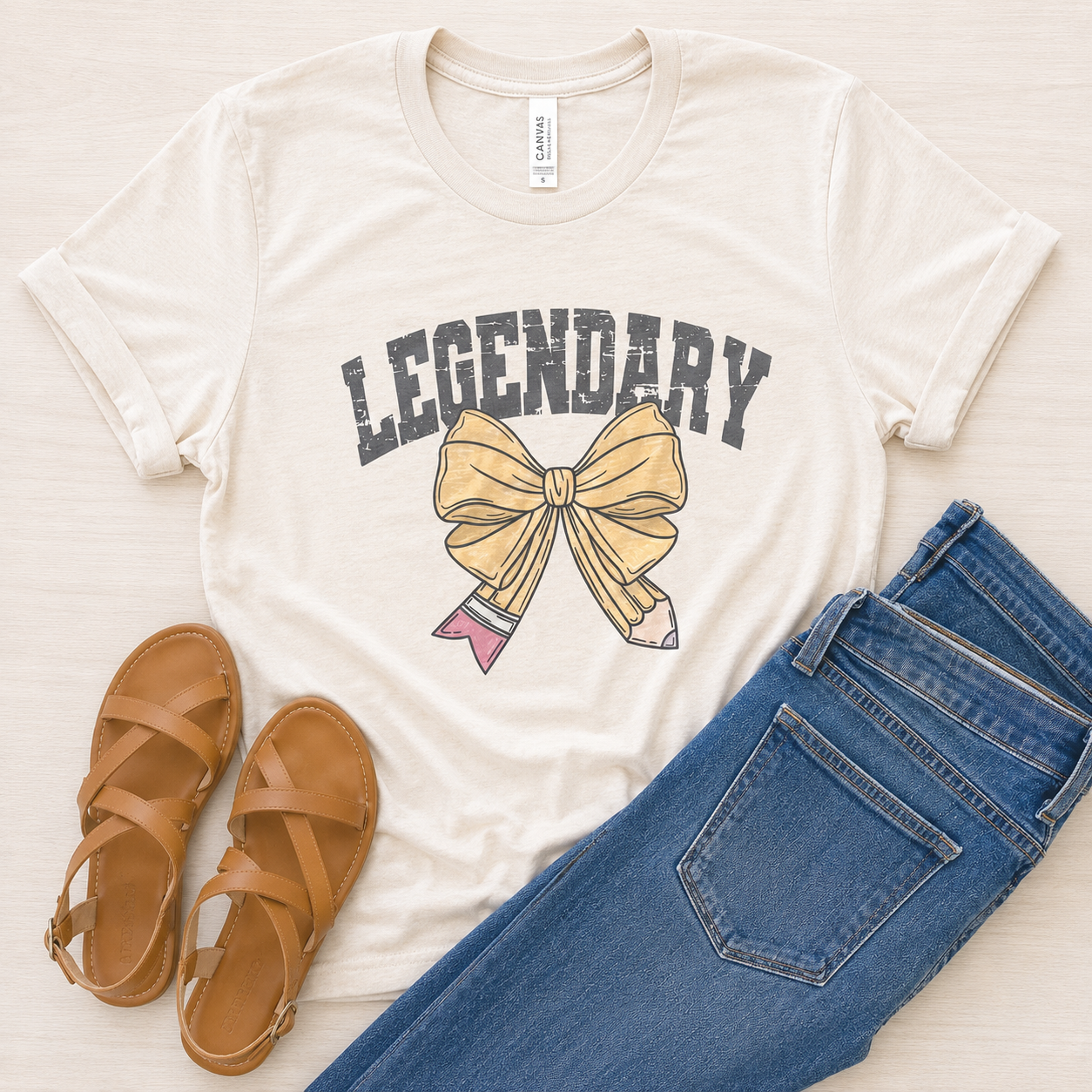 A cream-colored graphic t-shirt featuring the word 'LEGENDARY' in distressed dark gray letters above a yellow bow and pink pencil illustration. The t-shirt is styled in a flat lay with blue jeans and tan leather sandals on a light wood background.